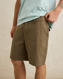 A man wearing olive brown linen shorts and a light blue t-shirt, standing against a neutral beige wall. Rhythm Men's Monterey Short - Fatigue