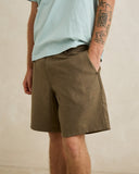 A person wearing olive brown textured shorts and a light blue t-shirt, standing against a neutral beige wall. Rhythm Men's Monterey Short - Fatigue