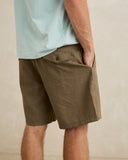 A close-up of a person wearing olive brown shorts with a light blue t-shirt, standing against a neutral beige background. Rhythm Men's Monterey Short - Fatigue