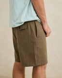A close-up of a person wearing olive brown textured shorts and a light blue t-shirt, standing against a neutral beige wall. Rhythm Men's Monterey Short - Fatigue