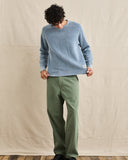 A model wearing a light blue knit crewneck sweater and olive green trousers, standing against a neutral beige fabric backdrop. Rhythm Mens Polu Crew Knit - Washed Blue