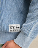 Close-up of a light blue textured knit sweater with a white sewn-in label that reads 'Rhythm Quality Made Garments' with a palm tree graphic, worn over a green shirt. Rhythm Mens Polu Crew Knit - Washed Blue