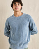A man wearing a light blue textured knit crewneck sweater, standing against a neutral beige fabric backdrop. Rhythm Mens Polu Crew Knit - Washed Blue