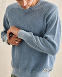 A close-up of a person wearing a light blue textured knit crewneck sweater, showing the ribbed collar, cuffs, and hem, with a small white brand patch near the lower hem. Rhythm Mens Polu Crew Knit - Washed Blue