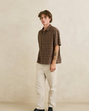 A man wearing a brown short-sleeve plaid button-up shirt and off-white trousers, standing against a textured beige studio wall. Rhythm Mens Relaxed Brady Check SS Shirt - Coffee