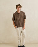 A man wearing a brown short-sleeve plaid button-up shirt and off-white trousers, standing against a textured beige wall. Rhythm Mens Relaxed Brady Check SS Shirt - Coffee