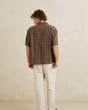 A man viewed from behind wearing a brown short-sleeve plaid button-up shirt and off-white trousers, standing against a textured beige wall. Rhythm Mens Relaxed Brady Check SS Shirt - Coffee