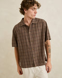 A man wearing a brown and beige plaid short-sleeve button-up shirt with white pants, standing against a textured beige background. Rhythm Mens Relaxed Brady Check SS Shirt - Coffee