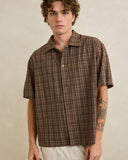 A man wearing a brown short-sleeve button-up shirt with a subtle plaid pattern, paired with off-white pants, standing against a neutral beige background. Rhythm Mens Relaxed Brady Check SS Shirt - Coffee