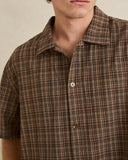 Close-up of a man wearing a brown short-sleeve plaid button-up shirt with a collar and white buttons, against a plain beige background. Rhythm Mens Relaxed Brady Check SS Shirt - Coffee