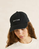 A woman wearing a black cotton baseball cap with the word 'RHYTHM' embroidered in white on the front, photographed against a plain light beige background. Rhythm Essentials Cap - Black