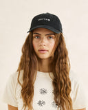 A woman with long wavy light brown hair wearing a black baseball cap with the word 'RHYTHM' embroidered in white on the front, paired with a cream-colored t-shirt with black floral line drawings, set against a plain beige background. Rhythm Essentials Cap - Black