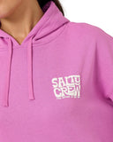 A close-up of a person wearing a bright pink hoodie with a white 'SALTY CREW' graphic printed on the left chest. Close up detail of the premium violet fleece fabric texture, crisp sleeve screen-printing, and Salty Crew internal neck branding.