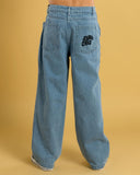 Wide-leg light blue denim jeans shown from the back, featuring a black embroidered 'Saint Cruz' logo on the right back pocket, worn by a model against a solid mustard yellow background. Back view of the Santa Cruz Yelling Stack Baggy Denim Jeans in Mid Blue, displaying the relaxed fit seat and pocket branding details.