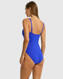 A woman modeling a bright royal blue one-piece swimsuit from the back, showing the open back design and thin shoulder straps, against a solid light gray background. Back view of the Sea Level Essentials Twist Front One Piece Swimsuit in Cobalt, displaying the smooth back coverage and adjustable straps.