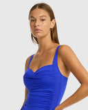 A woman modeling a bright royal blue ruched tankini swimsuit against a light gray studio background. Detailed close-up macro shot of the premium recycled eco fabric texture and twist-front details on the Cobalt Sea Level swimsuit.