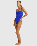 A woman modeling a bright royal blue one-piece swimsuit with ruched detailing on the bodice, photographed against a plain light gray studio background. Side profile view of the Cobalt Sea Level Essentials Twist Front Swimsuit, highlighting the sculpting fit and clean side seams.