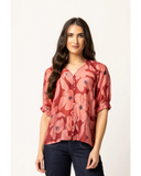 Detailed close-up macro view of the delicate Pink Bloom floral fabric print and premium stitching on the Seeking Lola Essence blouse. A woman modeling a short-sleeve button-up blouse with a dusty rose and coral floral print, paired with dark blue jeans, against a plain white studio background.
