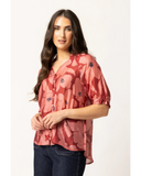 Side profile angle of the Seeking Lola Essence Top in Pink Bloom, highlighting the comfortable sleeve profile and relaxed body silhouette. A woman modeling a short-sleeve button-up blouse with a pink and red floral pattern, paired with dark blue jeans, against a plain white studio background.