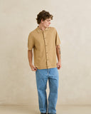 A man wearing a tan short-sleeve button-up shirt with a ribbed texture and light blue jeans, standing against a neutral beige textured wall. Rhythm Mens Textured Knit SS Shirt - Sand