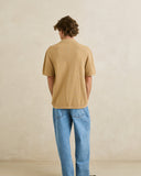A man viewed from behind wearing a short-sleeve textured knit t-shirt in a warm tan/beige color, paired with light blue jeans, standing against a neutral beige wall. Rhythm Mens Textured Knit SS Shirt - Sand