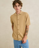A young man with curly brown hair wearing a short-sleeve, button-front knit shirt in a warm tan color, paired with blue jeans, standing against a neutral beige textured background. Rhythm Mens Textured Knit SS Shirt - Sand