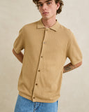 A man wearing a short-sleeve waffle-knit polo shirt in a warm tan/beige color, paired with blue jeans, standing against a neutral beige background. Rhythm Mens Textured Knit SS Shirt - Sand