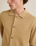 Close-up of a man wearing a short-sleeve waffle-knit polo shirt in a warm tan or camel color, showing the collar, front buttons, and textured fabric against a plain light beige background. Rhythm Mens Textured Knit SS Shirt - Sand