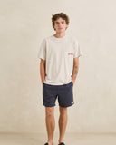 A man modeling a light beige crewneck pocket t-shirt with a red and blue chest graphic, paired with navy blue shorts, standing against a textured beige wall. Rhythm Mens Textured Linen Jam Shorts - Navy