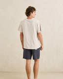 A man shown from the back wearing a light beige short-sleeve t-shirt and navy blue shorts, standing against a neutral textured beige wall. Rhythm Mens Textured Linen Jam Shorts - Navy