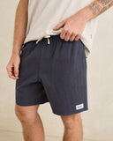 A man wearing navy blue drawstring shorts and a light beige t-shirt, standing against a neutral beige background. The brand name 'Mende' is visible on a small white label near the hem of the shorts. Rhythm Mens Textured Linen Jam Shorts - Navy