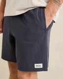 A close-up of a person wearing dark navy blue linen-blend shorts with a side pocket and a small white 'Rhythm.' label near the hem, paired with a light-colored t-shirt against a plain beige background. Rhythm Mens Textured Linen Jam Shorts - Navy