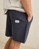 A close-up of a person wearing dark navy blue denim shorts with a side pocket and a white 'Rhythm.' brand label, paired with a light beige t-shirt against a neutral beige background. Rhythm Mens Textured Linen Jam Shorts - Navy