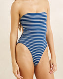 A woman wearing a strapless one-piece swimsuit with horizontal blue and white stripes, shown from the shoulders to the upper thighs against a plain light beige studio background. Front view of the Rhythm Vista Stripe Strapless One Piece in Deep Blue showing the bandeau neckline and horizontal stripe design.
