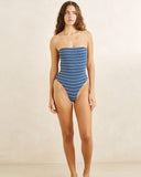 A woman modeling a strapless one-piece swimsuit with horizontal blue and white stripes, standing against a plain beige studio backdrop.