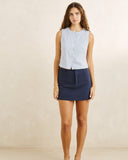 A woman stands against a plain beige studio wall wearing a sleeveless light blue and white vertically striped button-front top with a dark navy mini skirt. Rhythm Vista Stripe Vest Blue