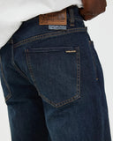 Close-up back view of dark blue Volcom jeans worn with a white shirt, showing the back pocket, stitching, and leather brand patch.