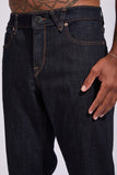 Close-up of a person wearing dark indigo denim jeans, showing the front waistband, button, belt loops, pocket, and hand resting on the thigh against a light gray background.