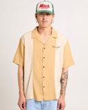 A man wearing a short-sleeved, button-down Wrangler shirt in beige and white colors with a left chest embroidered logo, paired with blue jeans and a red 'Mack' baseball cap. Front view of the Wrangler Bowlarama Short Sleeve Shirt in Wheat showing the relaxed boxy fit, camp collar, and button-up design.